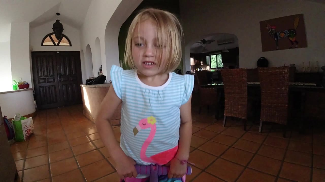 The young girl remains centered in the wide-angle frame, looking toward the lens with her hands low and tiled flooring stretching behind her.