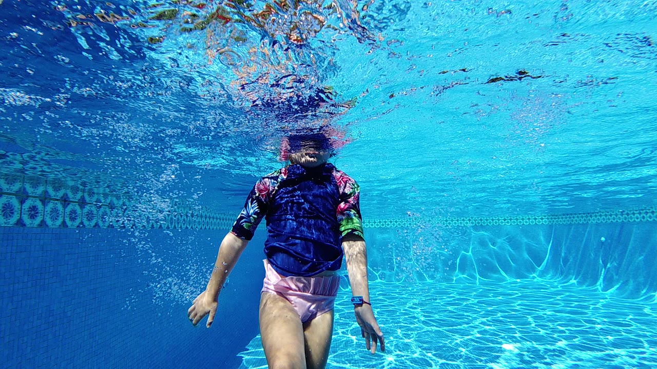 A young child in a blue rash guard stands underwater near the center of a bright blue swimming pool, with the surface rippling above.