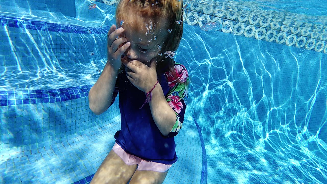 Underwater view of a child in a blue shirt and pink shorts near the pool surface; child's torso and head area visible with what appears to be swim goggles; blue mosaic tiles visible in background.