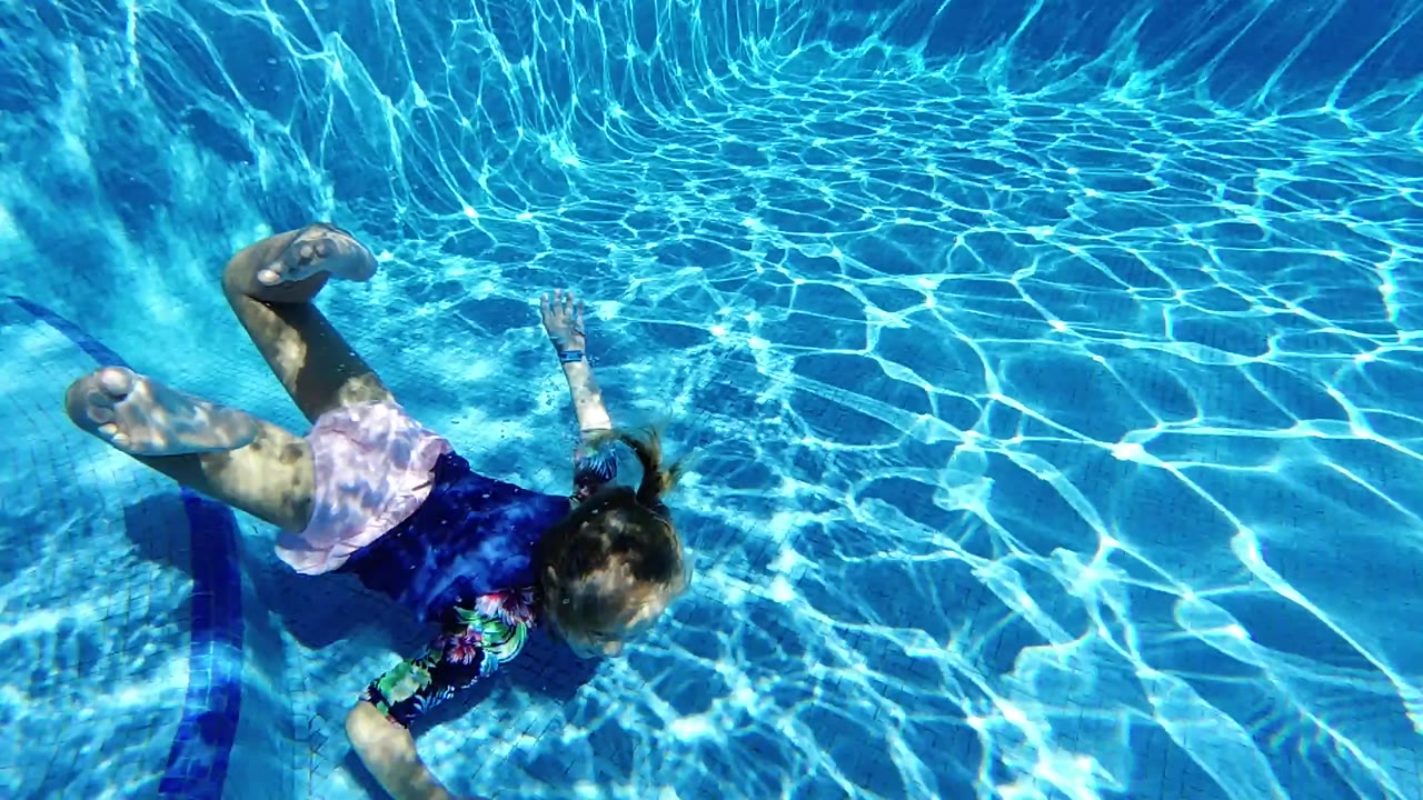 A young girl in a dark swim shirt swims underwater near the left pool wall, with bright blue water and rippled light across her back.