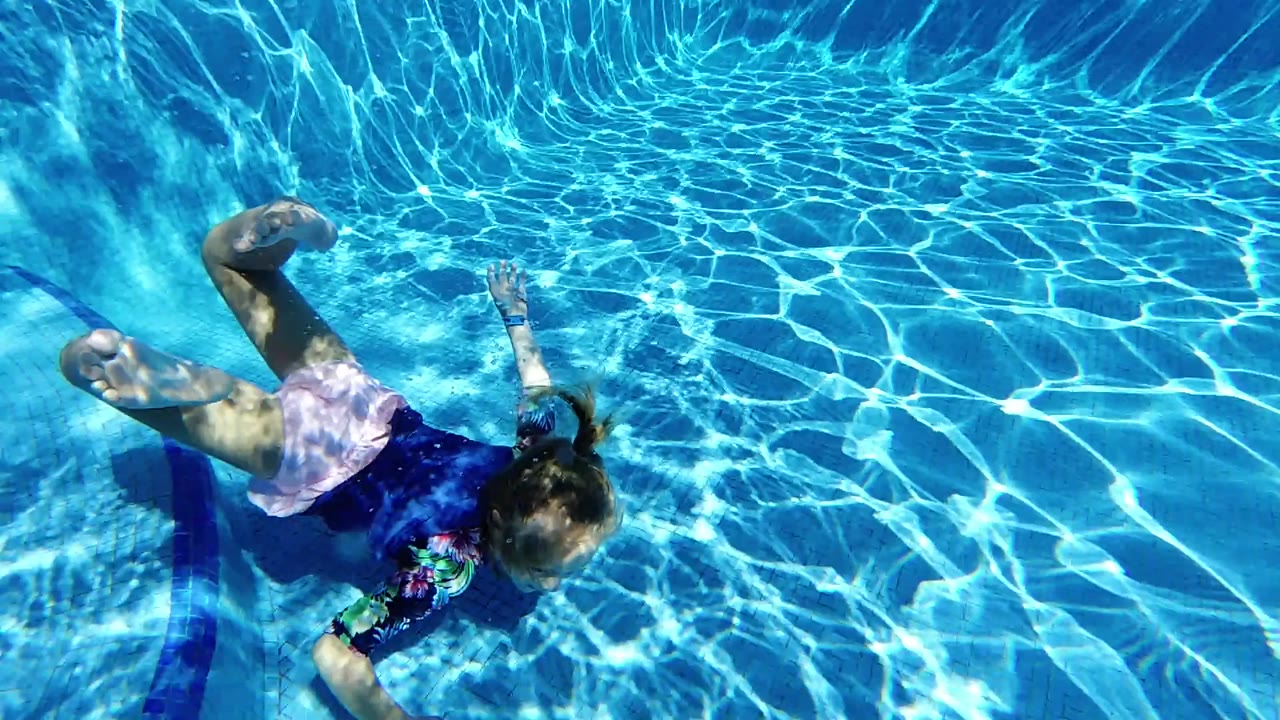 The girl glides underwater with one arm extended and bent legs, surrounded by blue pool water and high-contrast sunlight patterns.