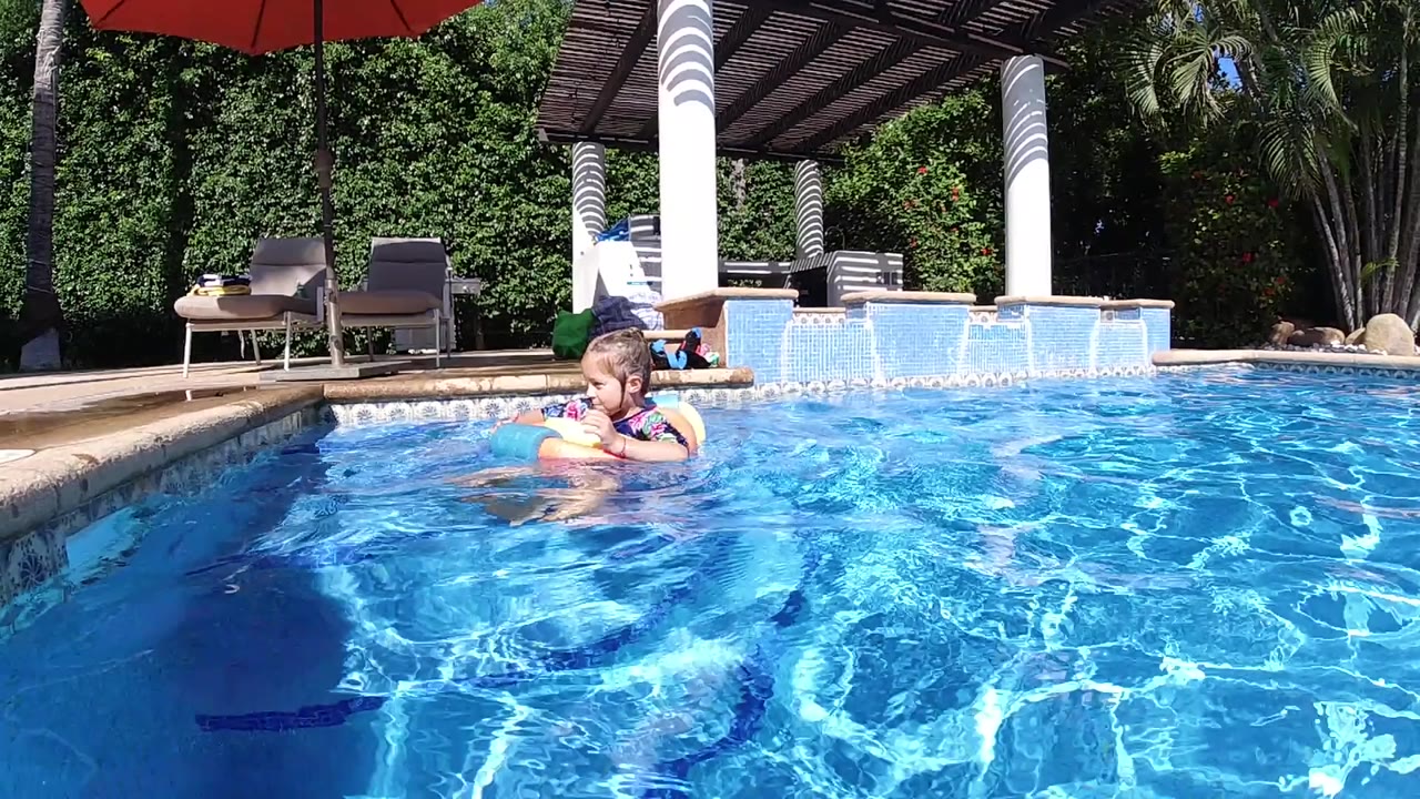 Young girl in a foam swim ring floats near the far side of a bright blue outdoor pool, with lounge chairs and dense greenery behind her.