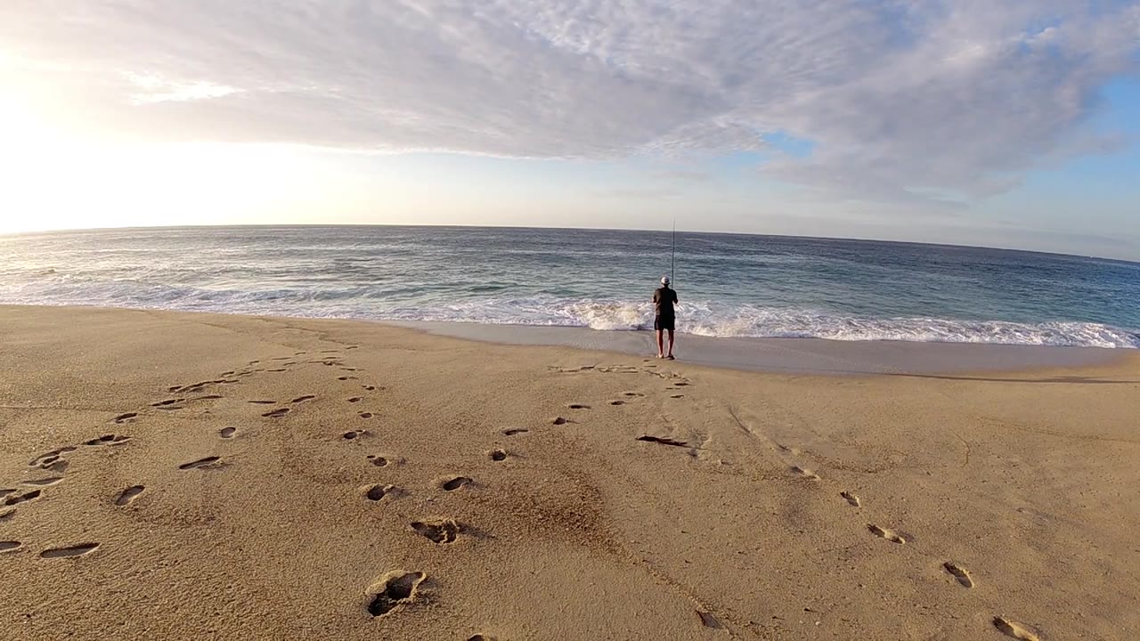 A single beach walker remains near the water as paired footprint trails mark the sand under a bright cloud bank.