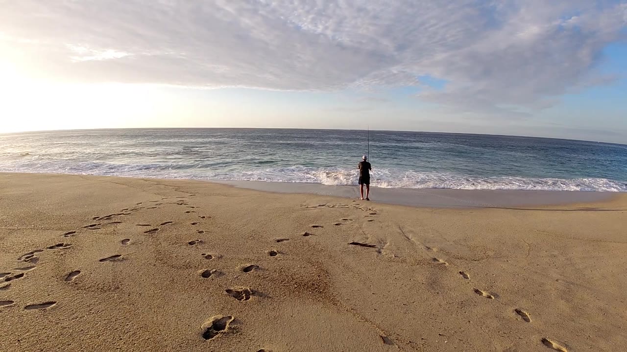 Footprints angle through the foreground while the lone figure stands closer to the breaking waves under layered clouds.