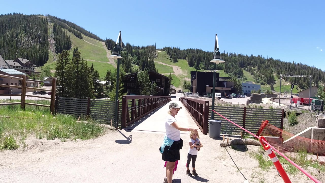 The woman and child remain near the bridge railing as the bright sun lights the gravel path, red barrier tape, lift equipment, buildings, and wooded hillside in the background.