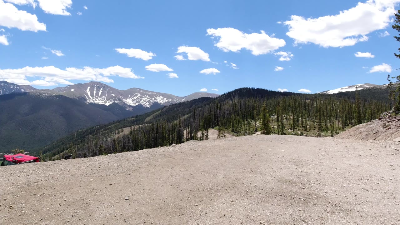 Wide Colorado Rockies overlook with a pale dirt foreground, forested ridges, snow-streaked mountains, blue sky, and a red car partly visible at the lower left with no visible people.
