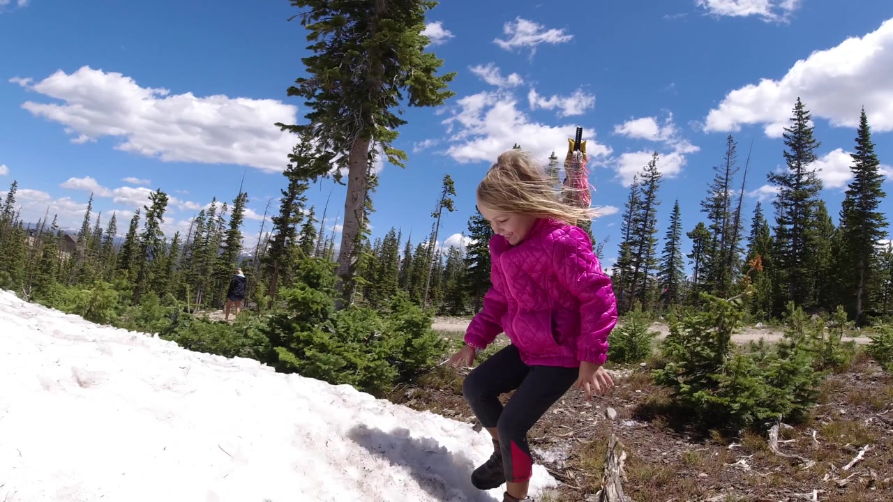 The girl passes closer to the snow patch, her pink jacket contrasting with the white snow and dark green conifers beyond.