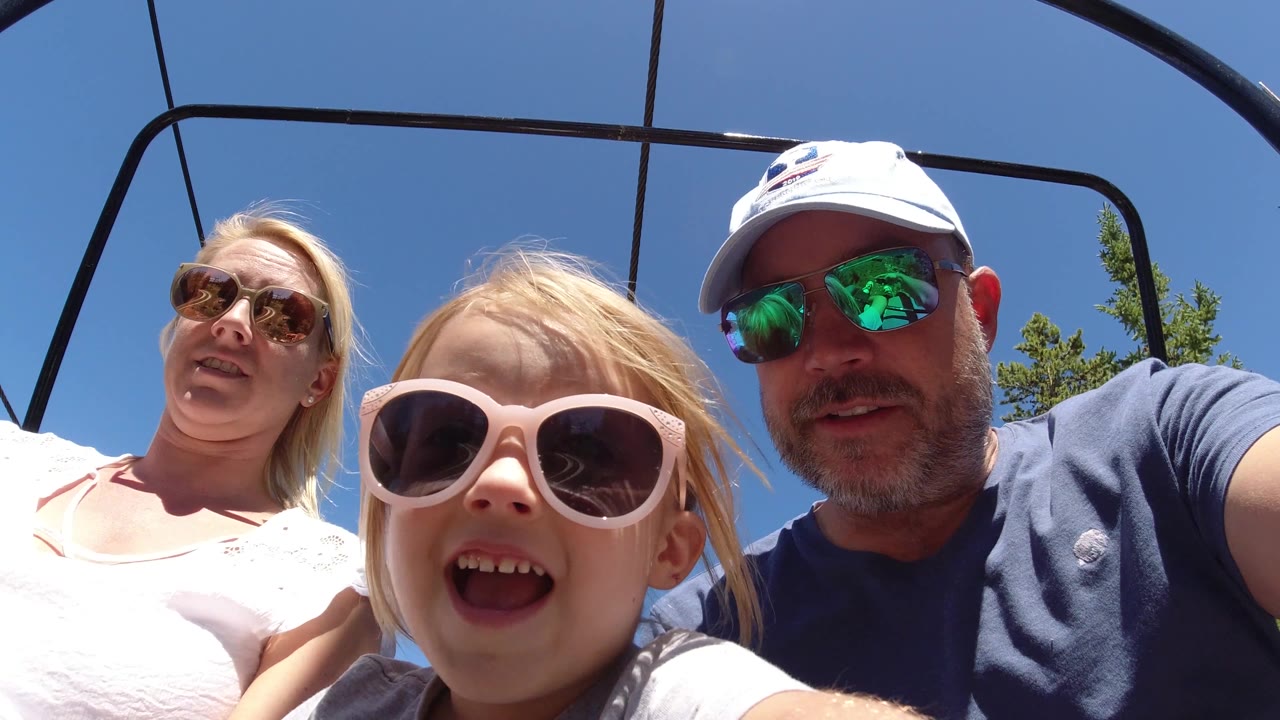 A blonde child in pale sunglasses sits close to the camera while two adults ride behind her on an open chairlift under a clear blue sky.
