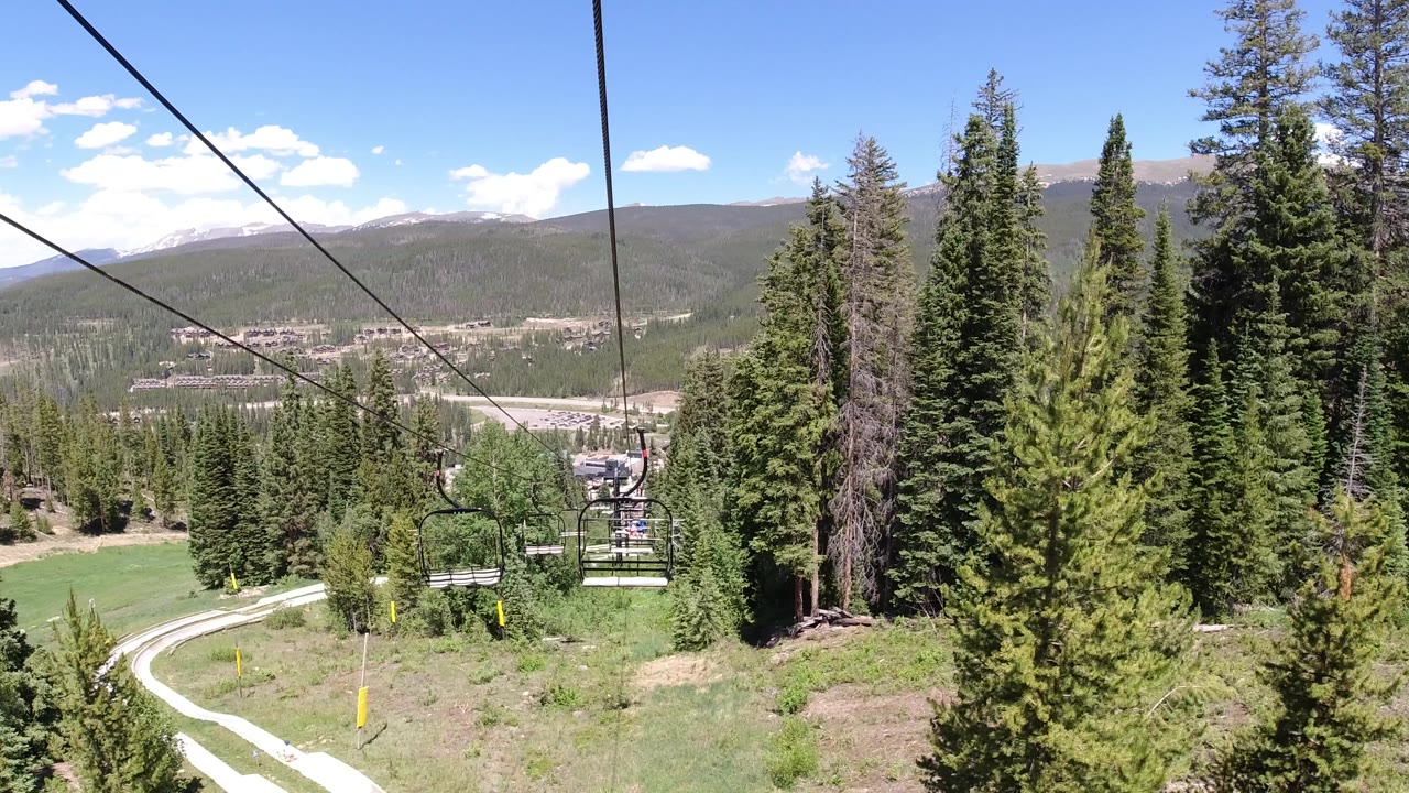 Opening view along chairlift cables above evergreen trees, with a winding road below, a town in the valley, mountain ridges, blue sky, and no visible people.