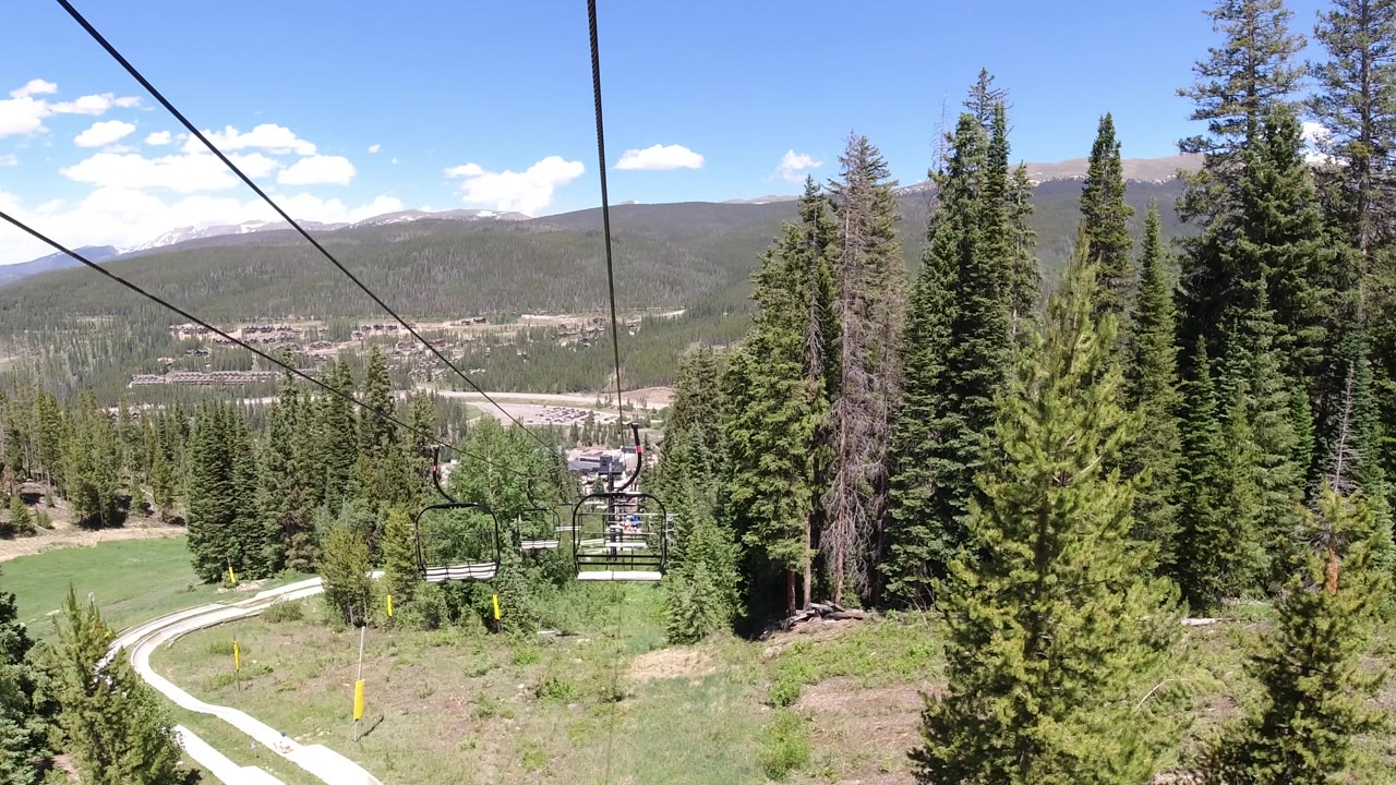 Forward-looking lift view across dense evergreens, with chairlift cables overhead, a pale winding road in the lower left, a valley town beyond, and no visible people.