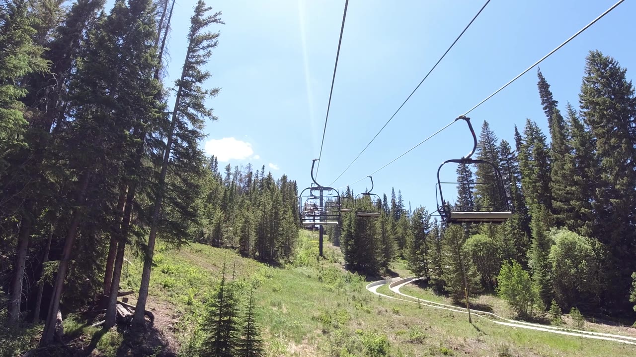 Looking up a ski lift line from the lower slope, an empty chair hangs from steel cables crossing diagonally over a green summer hillside with scattered conifer trees; no visible people.