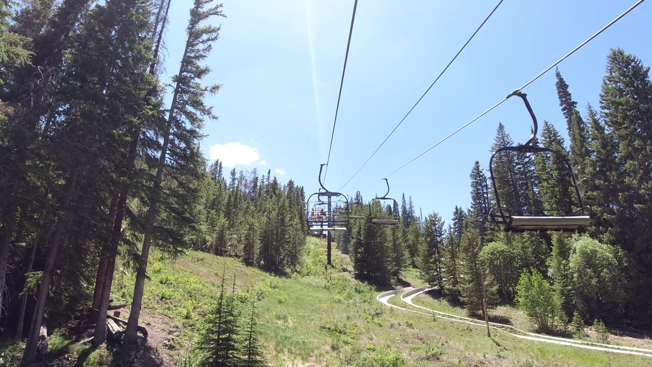 Two empty ski lift chairs are visible on the diagonal cable above a steep green summer hillside with conifer trees and bright blue sky; no visible people.