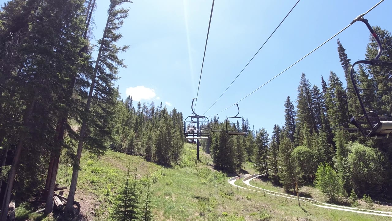 Looking up a ski lift line, empty chair on diagonal cable over green hillside with evergreen trees and partly cloudy sky.
