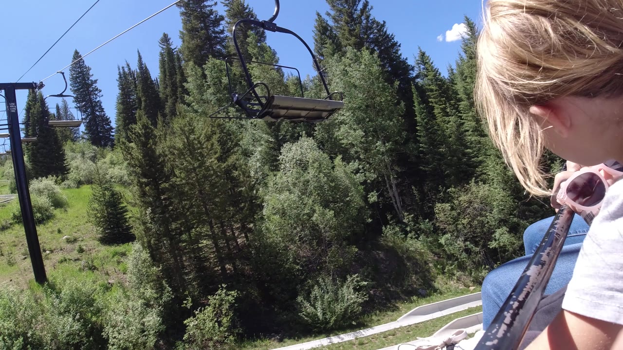 The child remains cropped at frame right as the chairlift looks out over dense green trees and a strip of pale path below.
