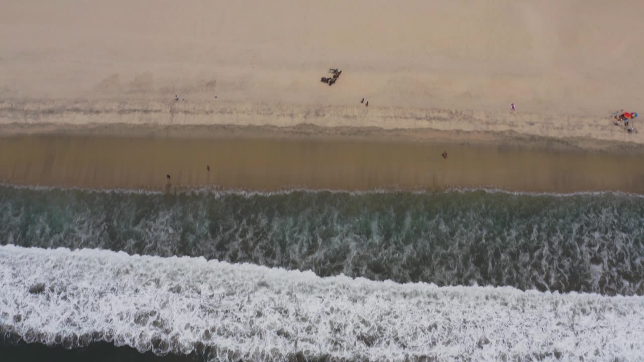 High overhead view of a Los Cabos beach with foamy surf crossing the lower frame, tan sand above it, and tiny people near the shoreline.