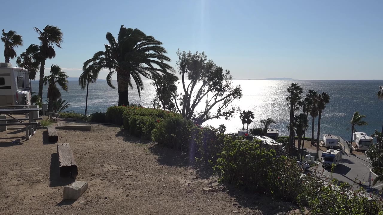 At 0:02, the Malibu view shows backlit palm crowns, a dry dirt foreground, dark shrubs, ocean glare, and small parked vehicles near the road below, with no visible people.