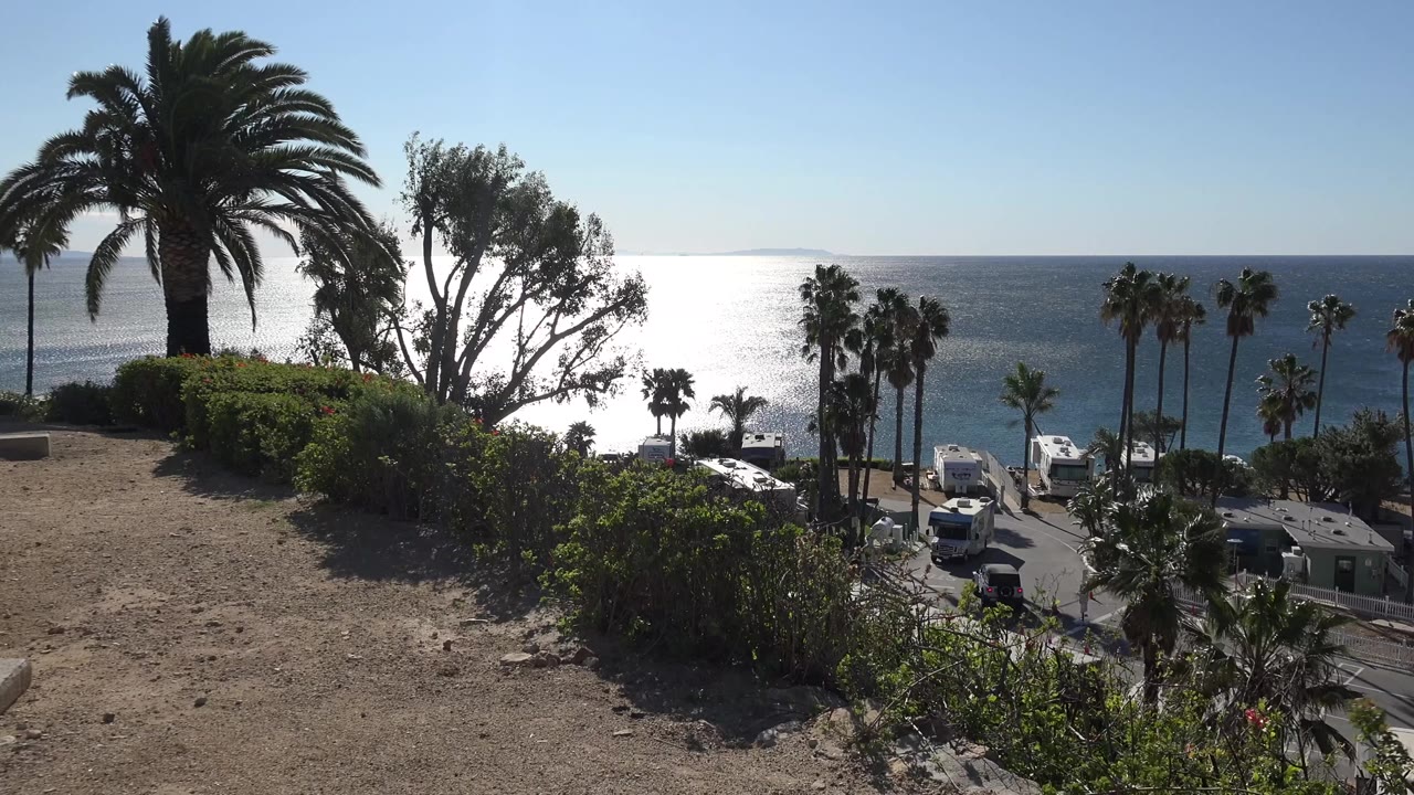 Opening frame of a Malibu coastal overlook with a dirt foreground, green shrubs, tall palms, bright ocean glare, parked vehicles below, and no visible people.
