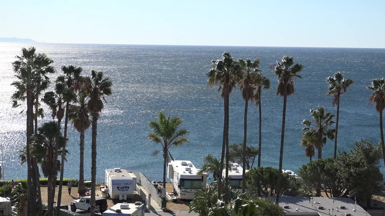 Opening frame of three white campers and tall palm trees above the Malibu oceanfront, with bright sun on the water and no visible people.