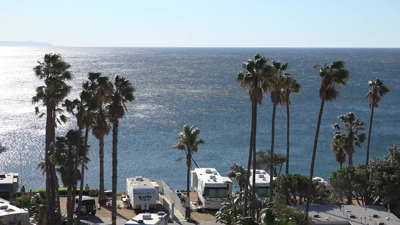 Opening frame of Malibu, California with tall palms above parked white RVs, bright glare on the ocean at left, a pale sky, foreground rooftops, and no visible people.