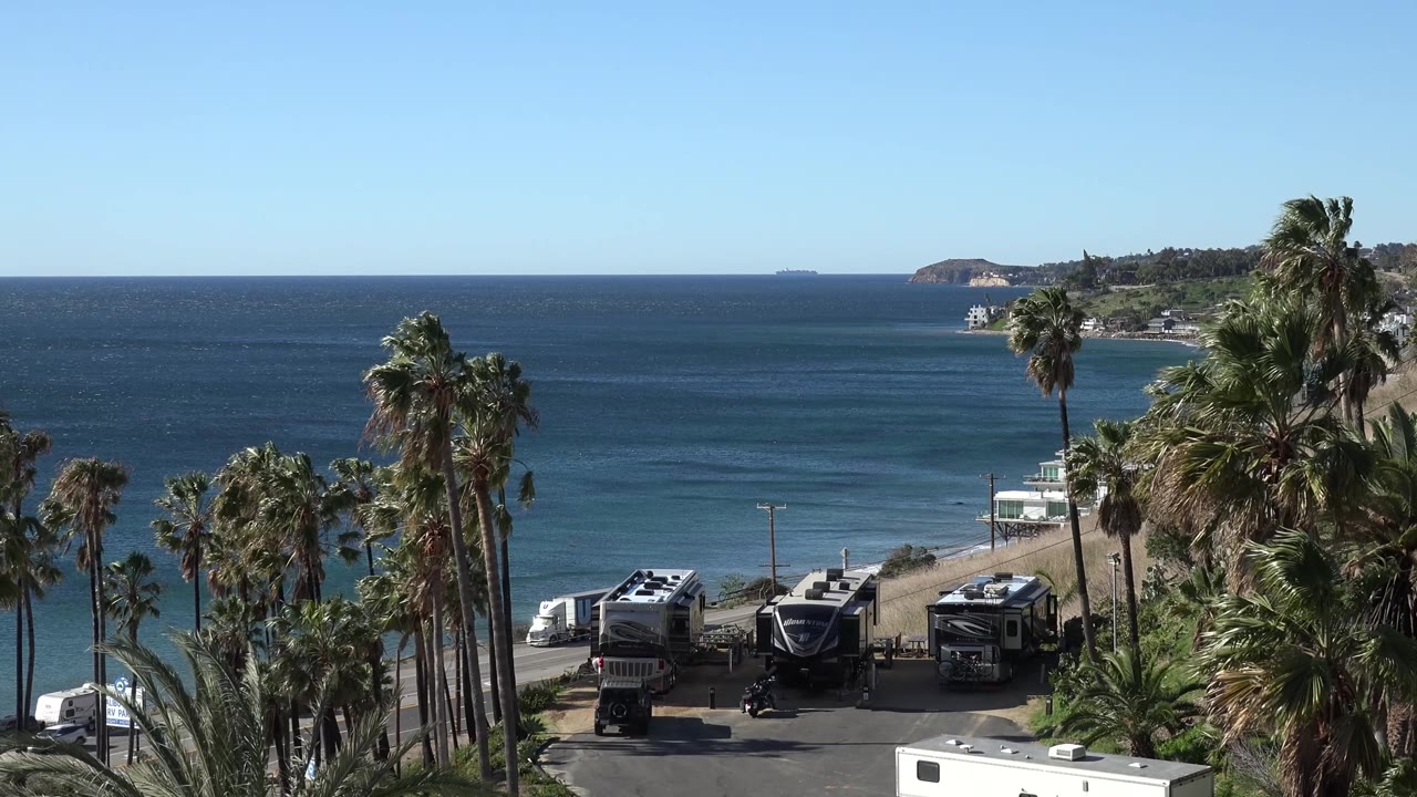 Malibu beachside parking view with palms crossing the foreground, several RVs below, ocean bands across the background, distant headland at right, and no visible people.
