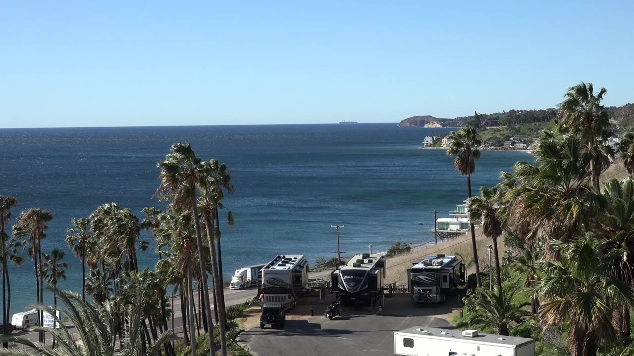 Opening Malibu frame with tall palms at left and right, parked RVs below, blue ocean beyond, a distant headland on the horizon, and no visible people.