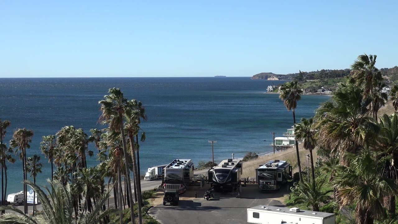 Mid-clip Malibu frame with the same elevated view, RV roofs visible below the palms, darker blue water offshore, a distant landform, and no visible people.