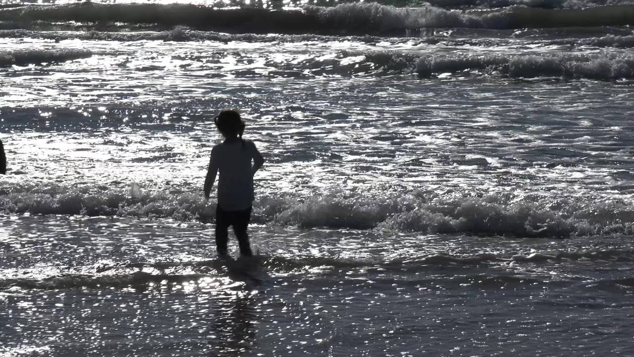 A silhouetted young girl stands in shallow ocean water at Malibu, California, with foamy waves and bright reflected light behind her.