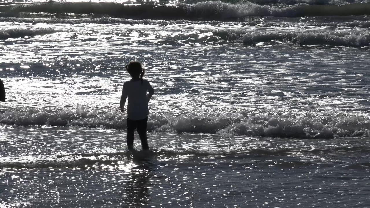 The young girl remains silhouetted in the Malibu surf as a low wave breaks across the wet sand in front of her.