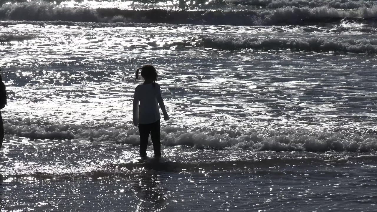 A young girl stands near the waterline at Malibu, California, separated from the viewer by wet sand and backed by bright, choppy surf.
