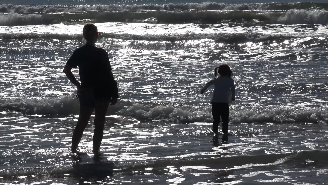 At 0:00 in Malibu, California, a silhouetted woman stands at the left while a child faces the incoming surf near the center of the frame.
