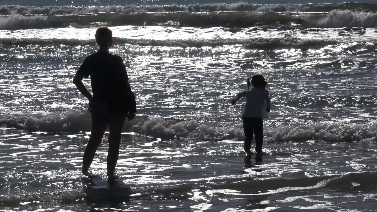 At 0:01 in Malibu, California, the adult remains in the left foreground while the child stands farther out as a low wave breaks around their legs.