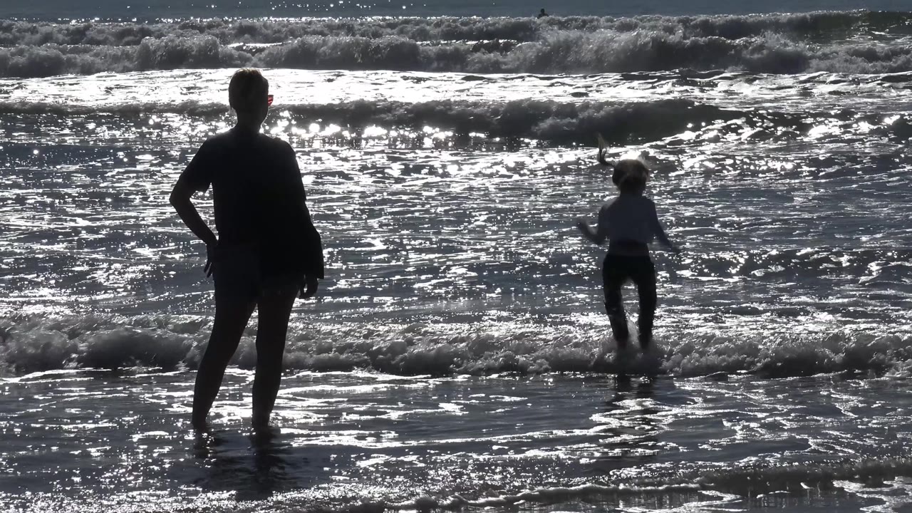 At 0:02 in Malibu, California, the child moves through a small breaker on the right while the adult silhouette stands planted on the left.