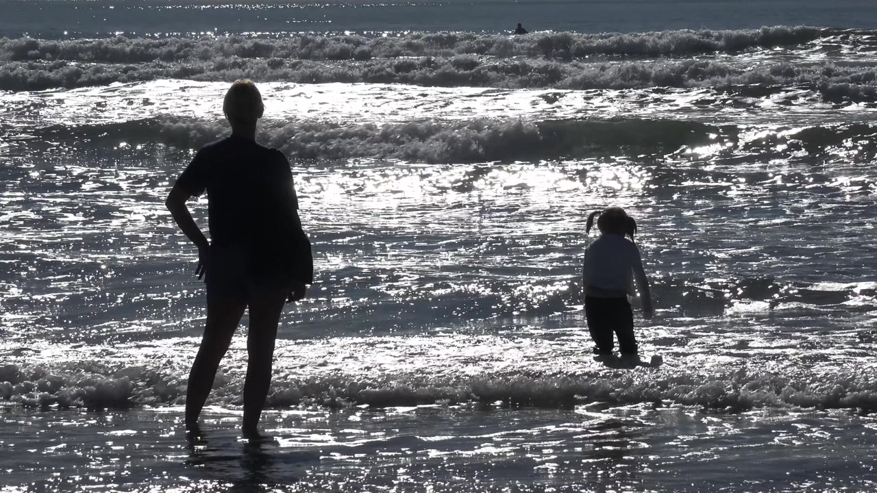 Silhouetted adult and child stand separated in shallow ocean water.