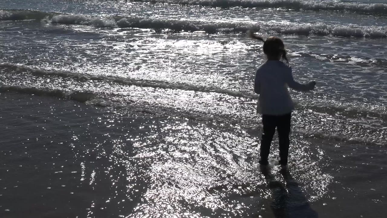 A young girl stands near the Malibu shoreline in shallow water as the low sun reflects across wet sand and small waves break behind her.