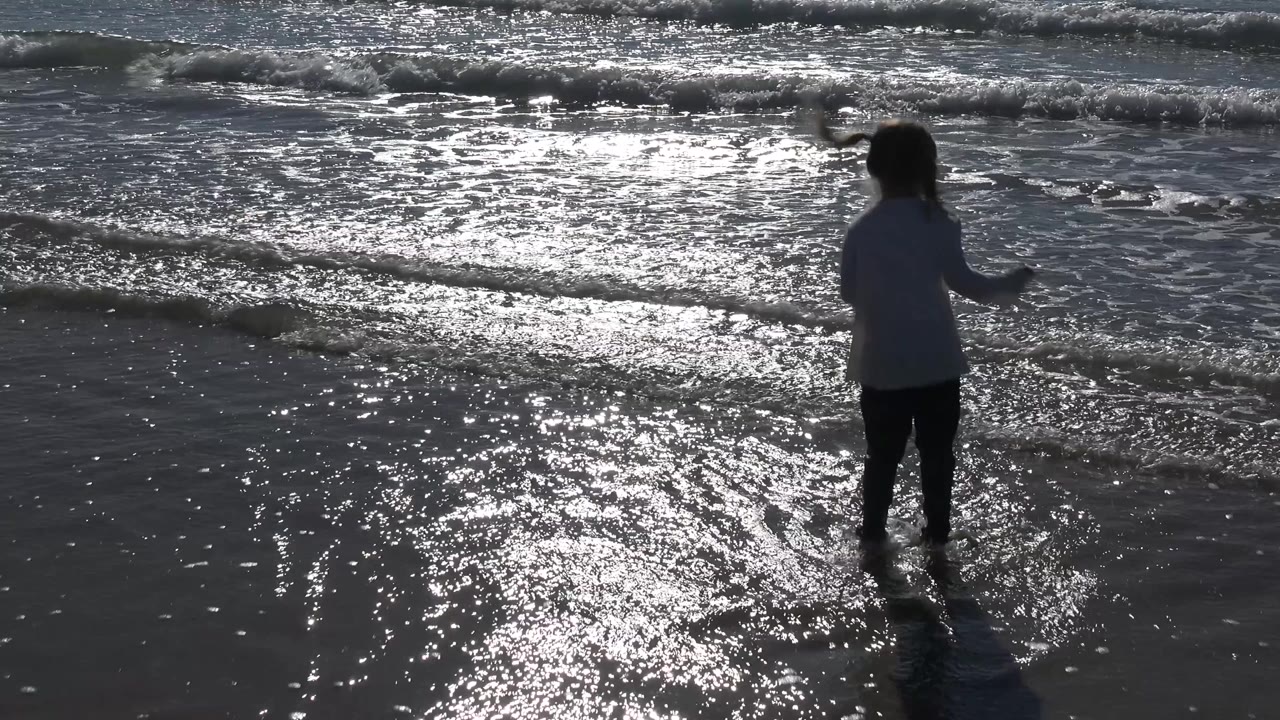 Young girl facing the water as a small wave approaches.