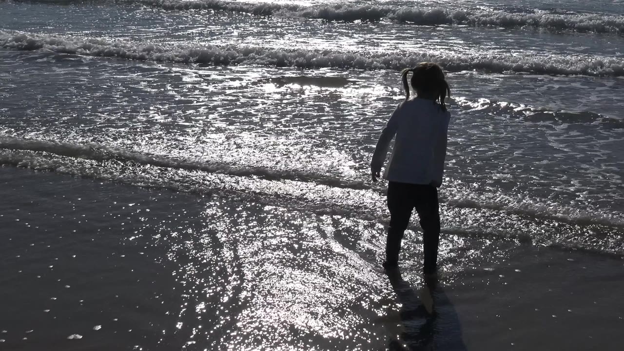 A backlit girl with a visible ponytail stands near the Malibu waterline as foam gathers across the background waves and glare fills the sand.