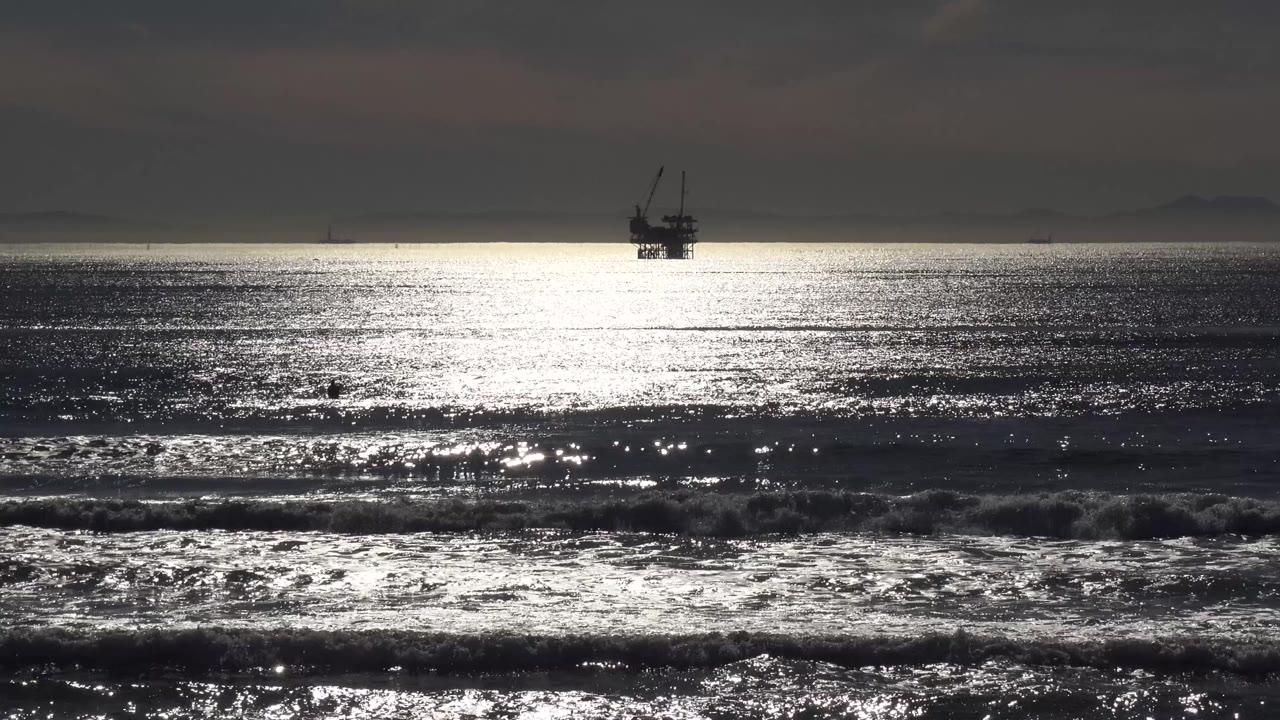 Dark ocean surf with bright sunset reflection and an offshore platform on the horizon.