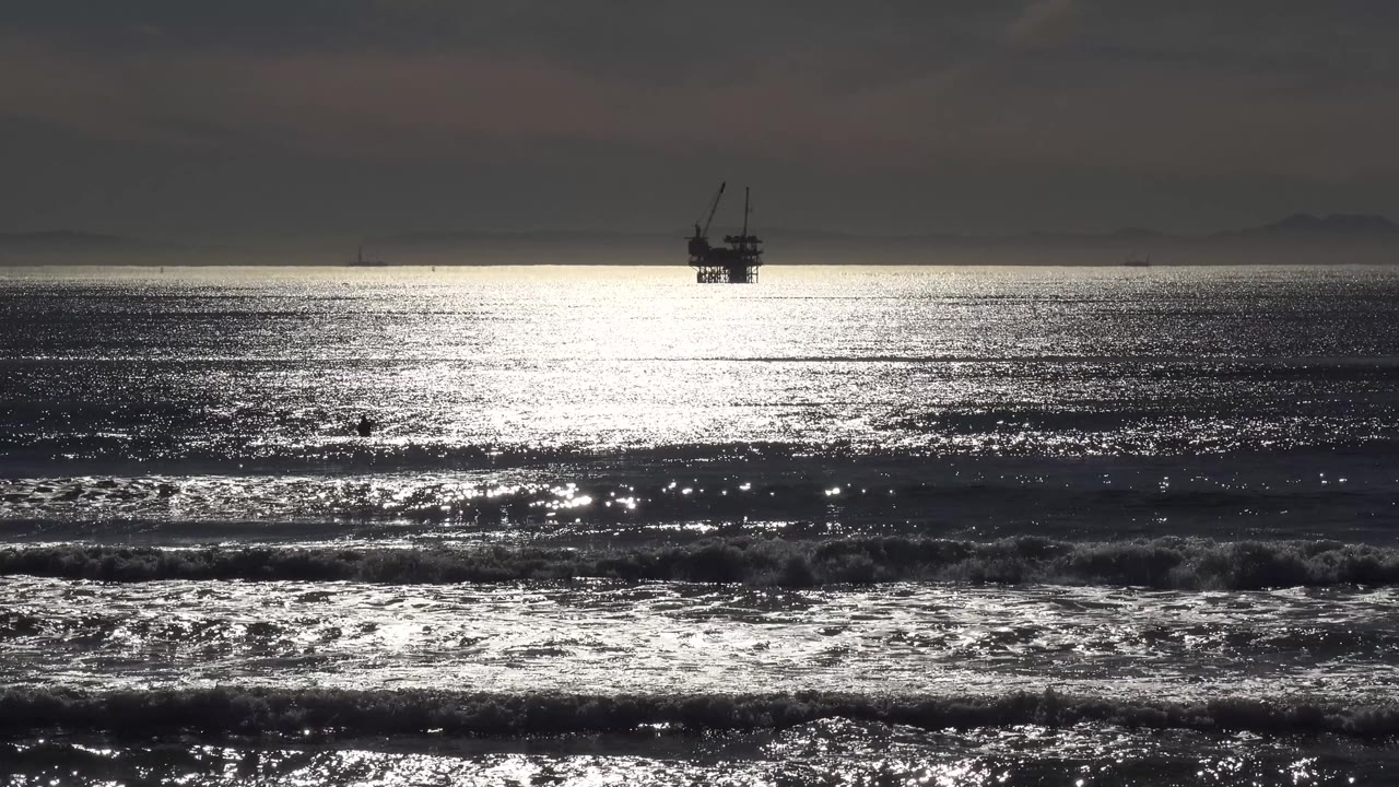 A Malibu sunset view shows the offshore platform centered against low clouds while broken surf lines catch bright reflected light, with no visible people.