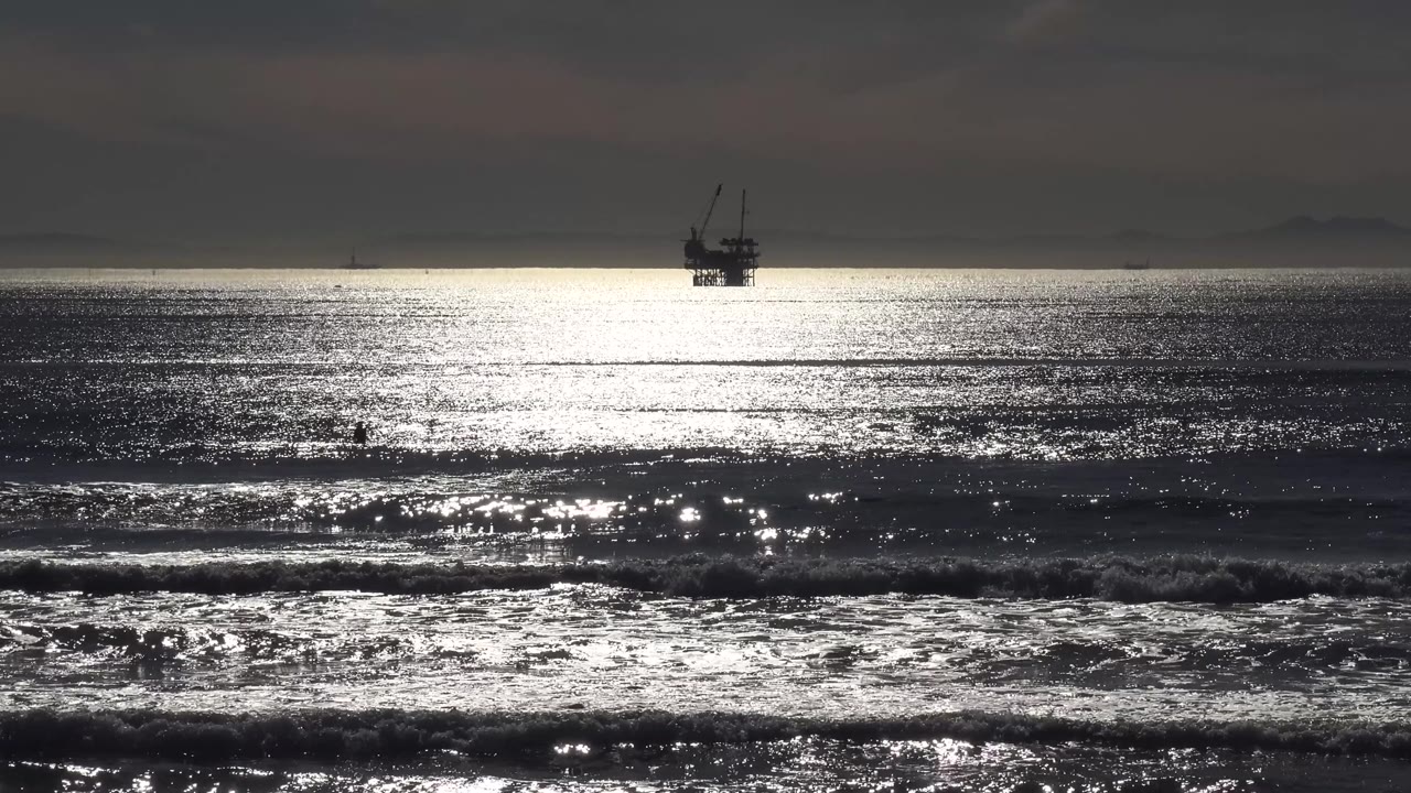 The Malibu horizon sits under a dark cloud layer as glare spreads across the ocean surface and a distant offshore platform remains visible, with no visible people.