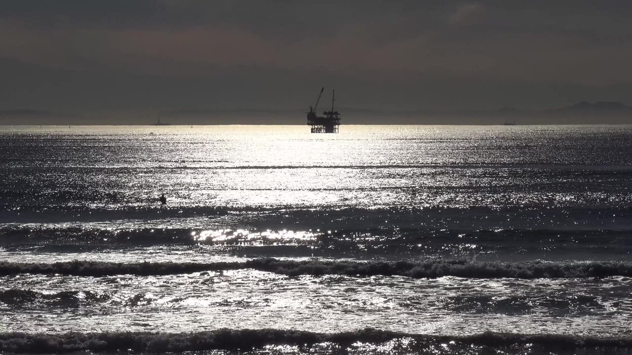 Malibu surf breaks across the foreground while the offshore platform appears as a small dark structure beyond the bright water, with no visible people.