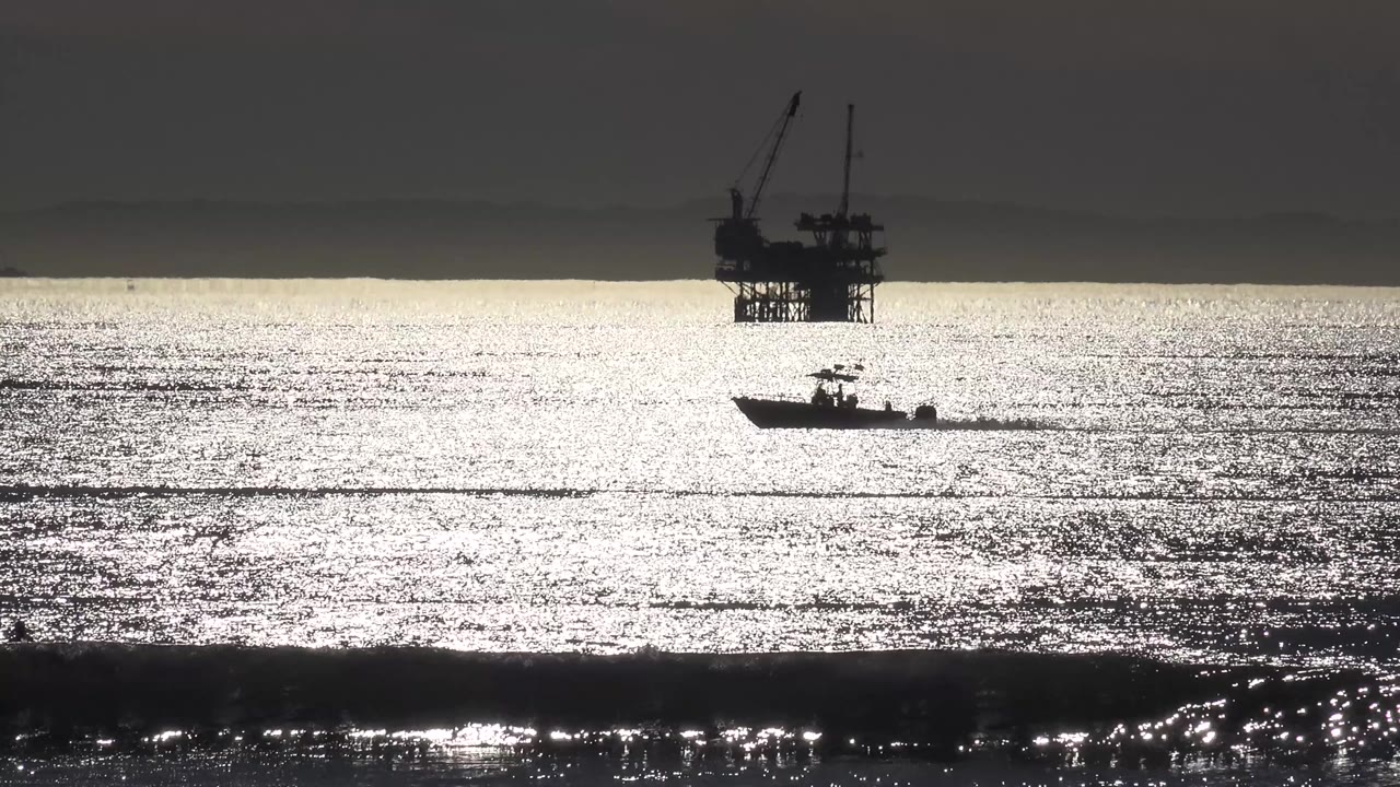 At 0:01 in Malibu, California, the small boat continues across the reflective ocean while the oil platform remains high in the background and no visible people appear.