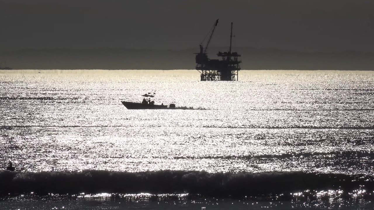 At 0:02 in Malibu, California, the boat sits farther right against dense silver water texture, with the oil rig silhouetted above the horizon and no visible people.