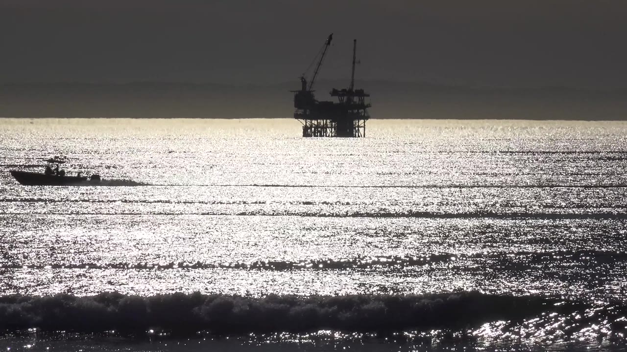 At 0:03 in Malibu, California, the boat is near the left side of the frame as white glare covers the ocean and the oil platform stands in silhouette beyond it, with no visible people.