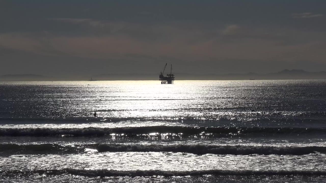 Malibu, California ocean view at 0:00 with a distant large boat near the horizon, bright silver reflection on dark water, low clouds, breaking surf, and no identifiable people visible.