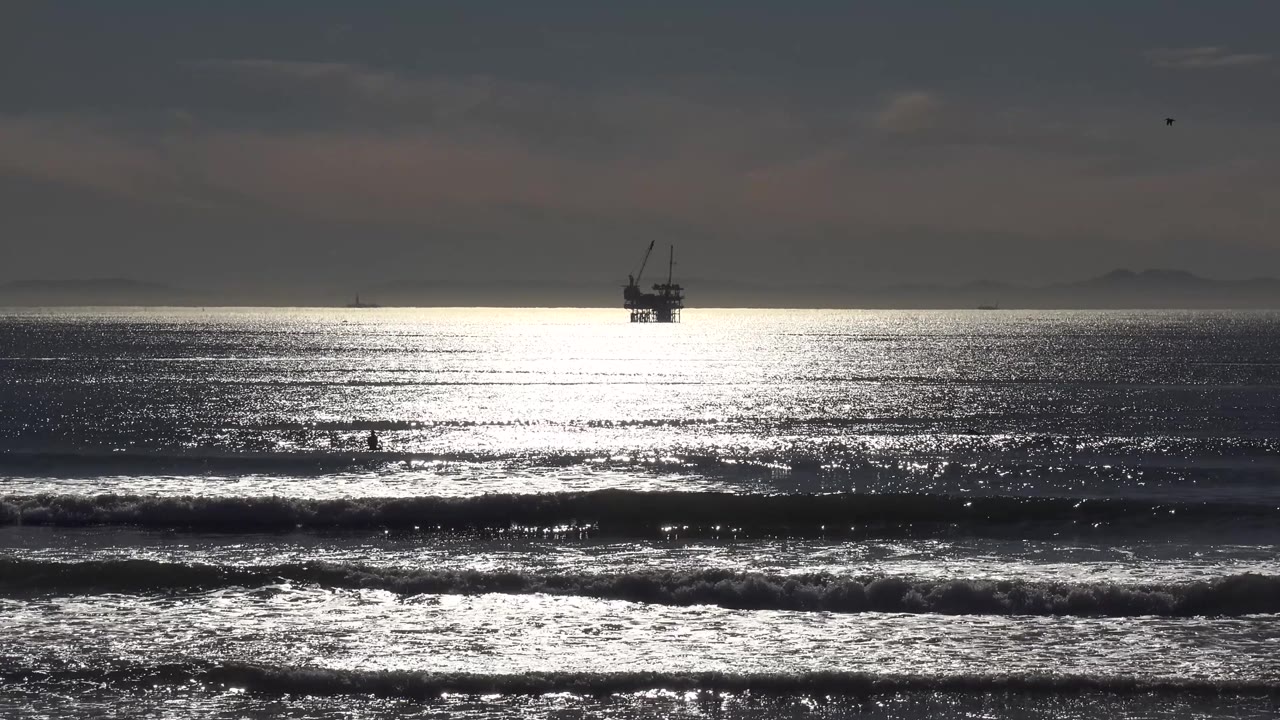 Malibu, California ocean frame at 0:02 places the distant boat toward the right of the bright reflection, with rolling surf below, a shadowed sky above, and no identifiable people visible.