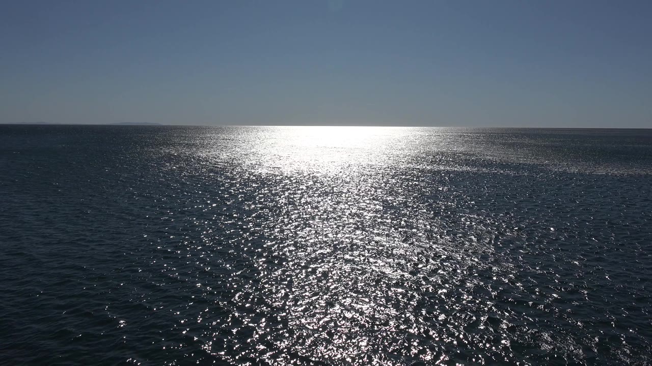 A broad Malibu ocean view shows the sun glare stretching across textured water slightly left of center under a cloudless sky, with no visible people.