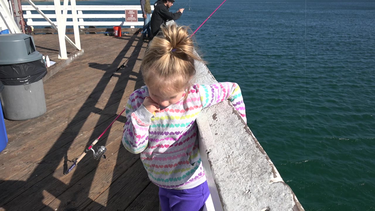 A young girl in a patterned sweatshirt stands at a white railing over the ocean in Malibu, California, with fishing rods and background people on the pier.