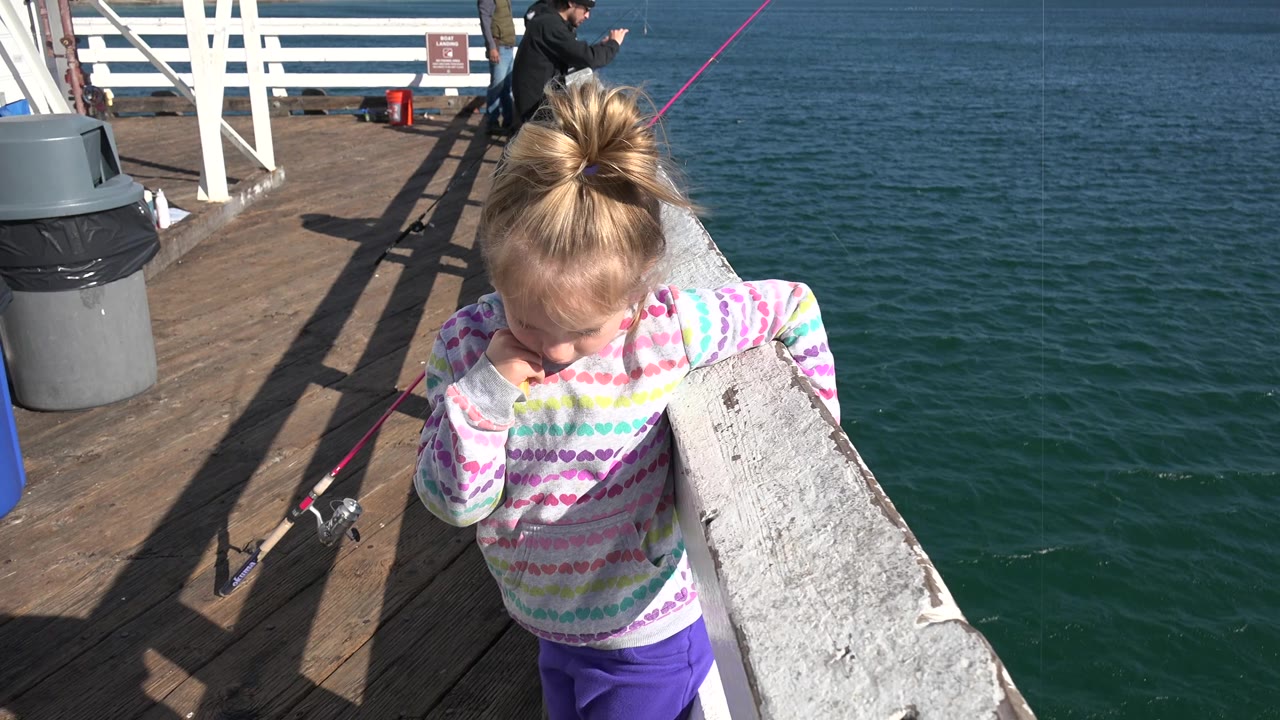The child remains near the white railing in Malibu, California, looking down as sun hits the pier deck, with ocean water and fishing rods behind her.