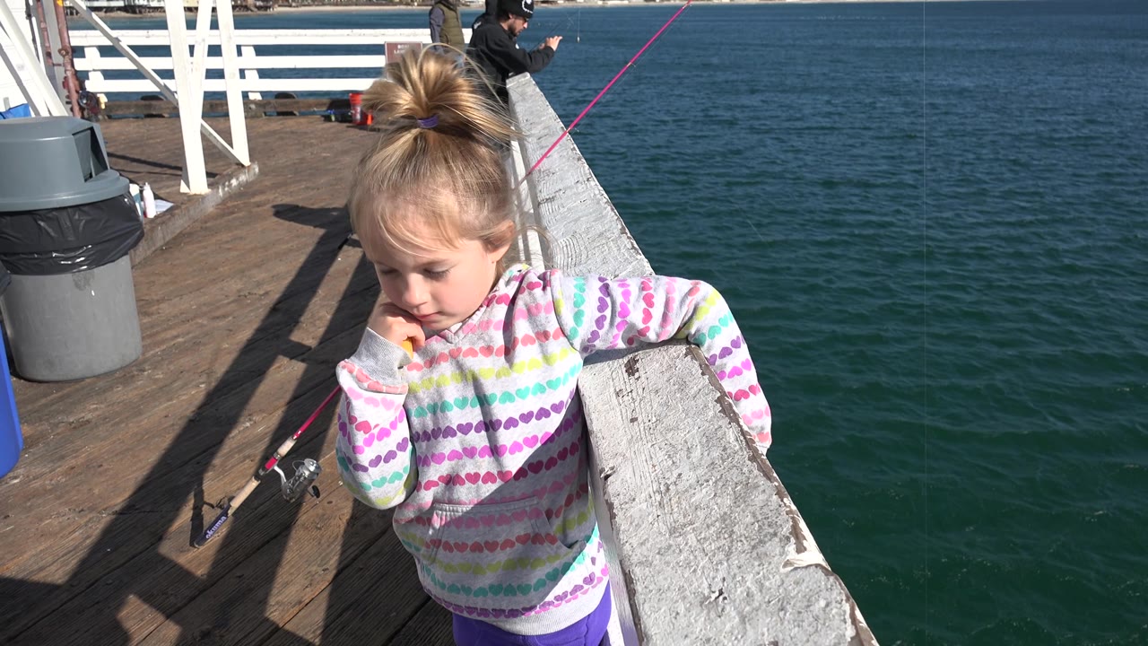 The girl turns partly toward the camera at the Malibu railing, with dark blue ocean water to the right and fishing activity visible farther back on the pier.