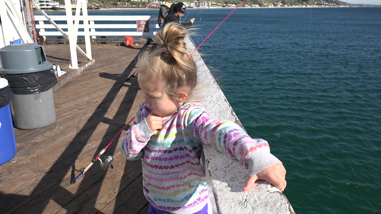 A young girl in a patterned hoodie holds a white boat rail on a Malibu boat, with fishing rods, bins, deck boards, open water, and an adult in the background.